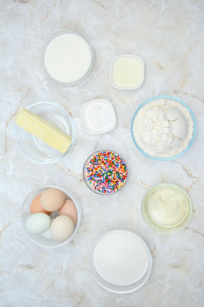 Flat lay shot of the ingredients for making the Funfetti Cake Roll. All are placed in separate glass bowls, and are on a marble surface.