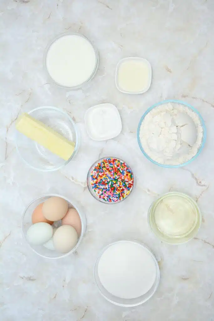Easy From Scratch Confetti Birthday Cake Roll Recipe with Cream Cheese Frosting 9 Flat lay shot of the ingredients for making the Funfetti Cake Roll. All are placed in separate glass bowls, and are on a marble surface.