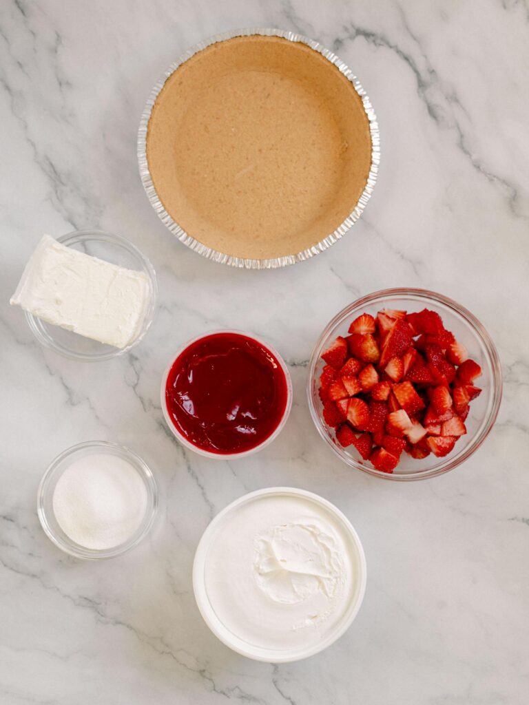 Best No-Bake Strawberry Cream Cheese Pie 7 Flat lay shot of the ingredients to make The Best No-Bake Strawberry Cream Cheese Pie, such as chopped fresh strawberries, gelatin, a tub of cool whip, strawberry jam, a block of cream cheese in a glass bowl and store-bought pie crust.