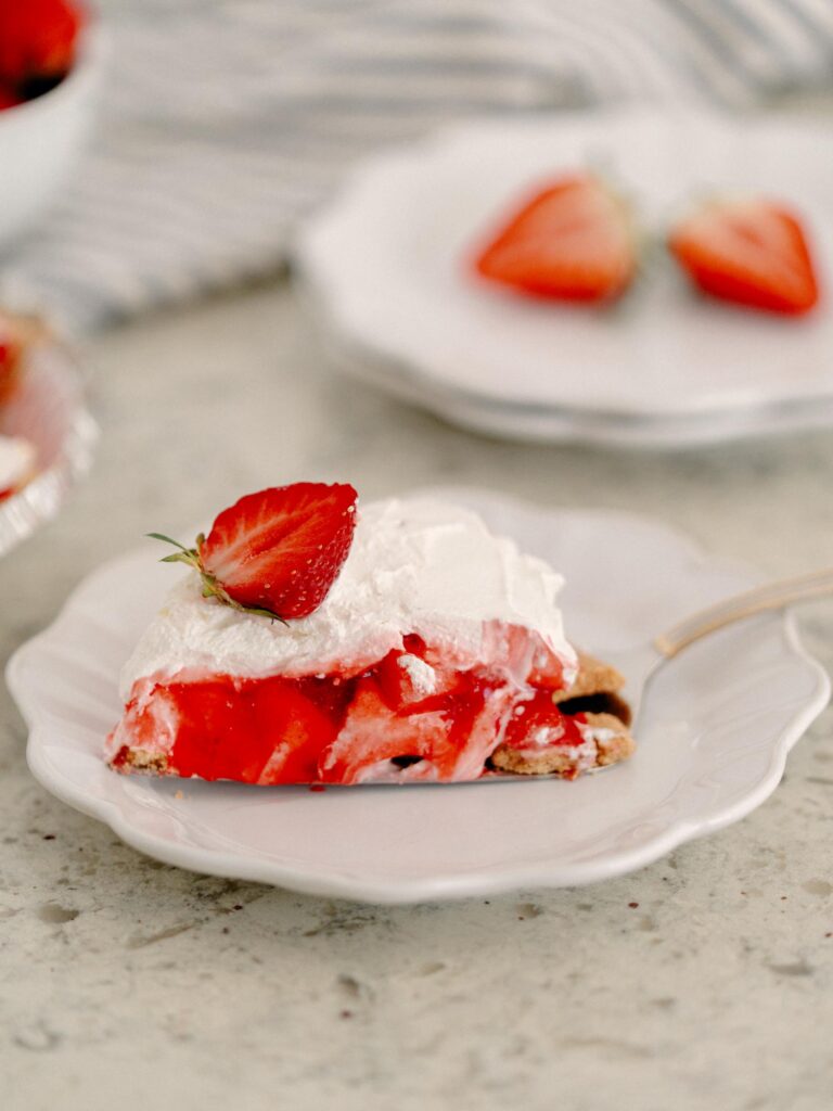 Best No-Bake Strawberry Cream Cheese Pie 5 Focus shot on a slice The Best No-Bake Strawberry Cream Cheese Pie on a white dessert plate. The pie slice is topped with half a fresh strawberry.