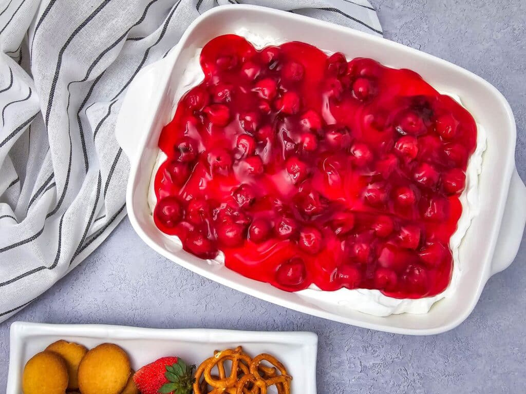 Flat lay shot of a white baking dish with the finished 4-Ingredient Cherry Cheesecake Dip. Beside the baking dish is a white rectangular platter with assorted dippables such as butter cookies, strawberries, mini pretzels.