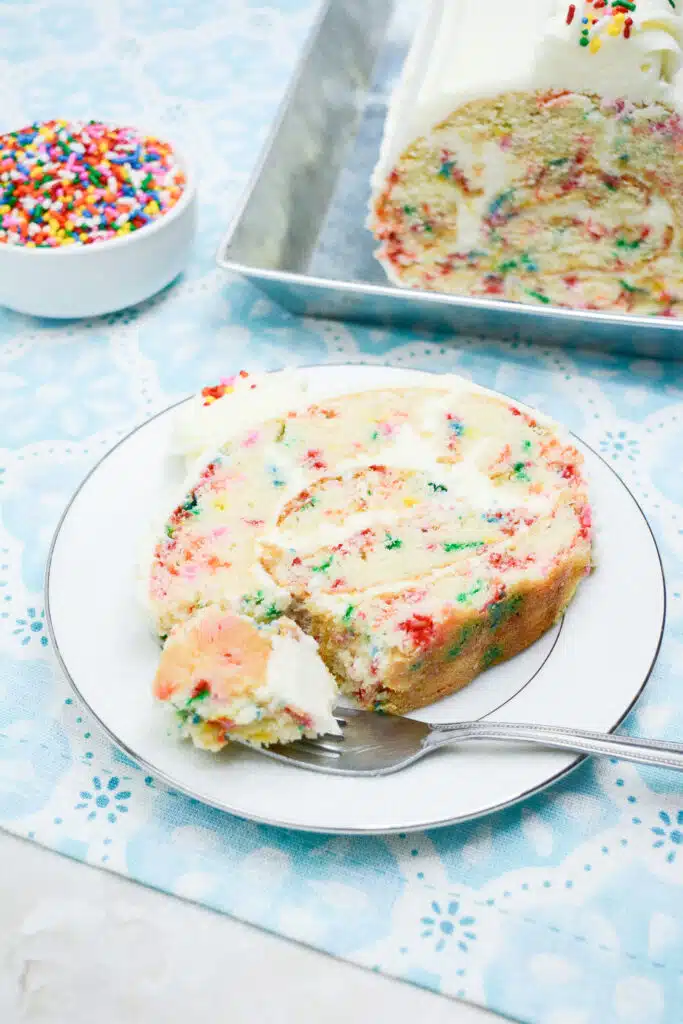 Easy From Scratch Confetti Birthday Cake Roll Recipe with Cream Cheese Frosting 8 A slice of the Delightful Funfetti Birthday Cake on a white dessert plate with a fork that has cut through a piece of the cake.