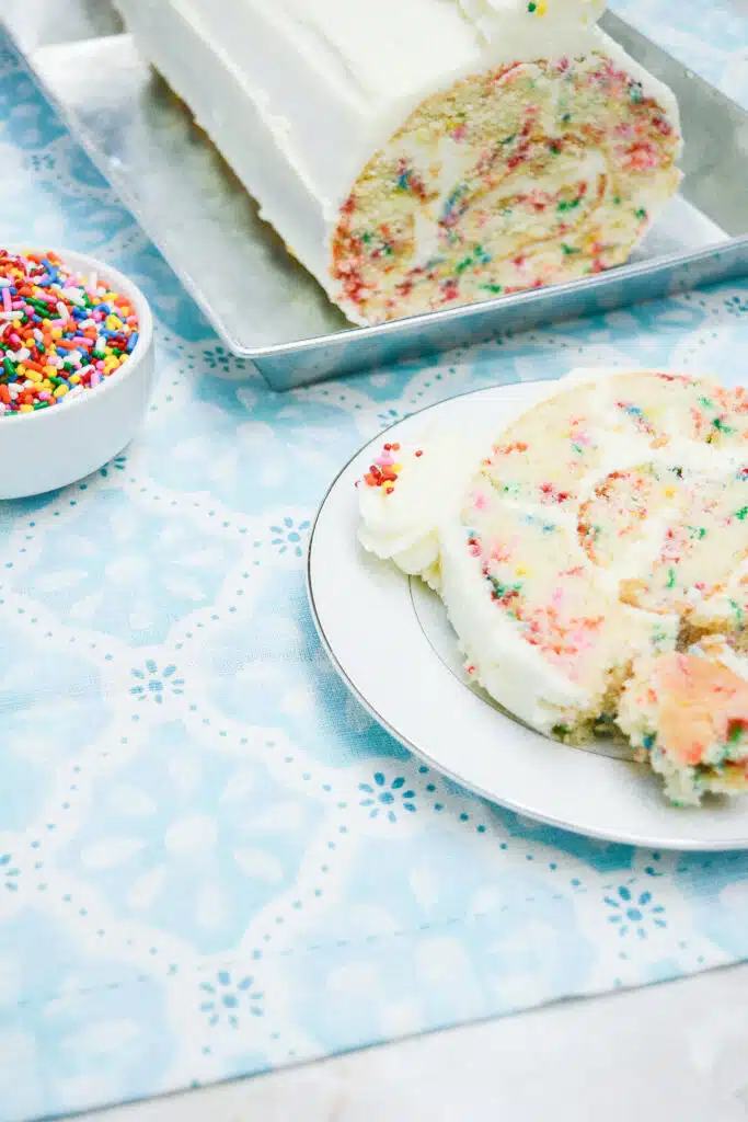 Easy From Scratch Confetti Birthday Cake Roll Recipe with Cream Cheese Frosting 5 A partial shot of the Delightful Funfetti Birthday Cake Roll slice on a white dessert plate.