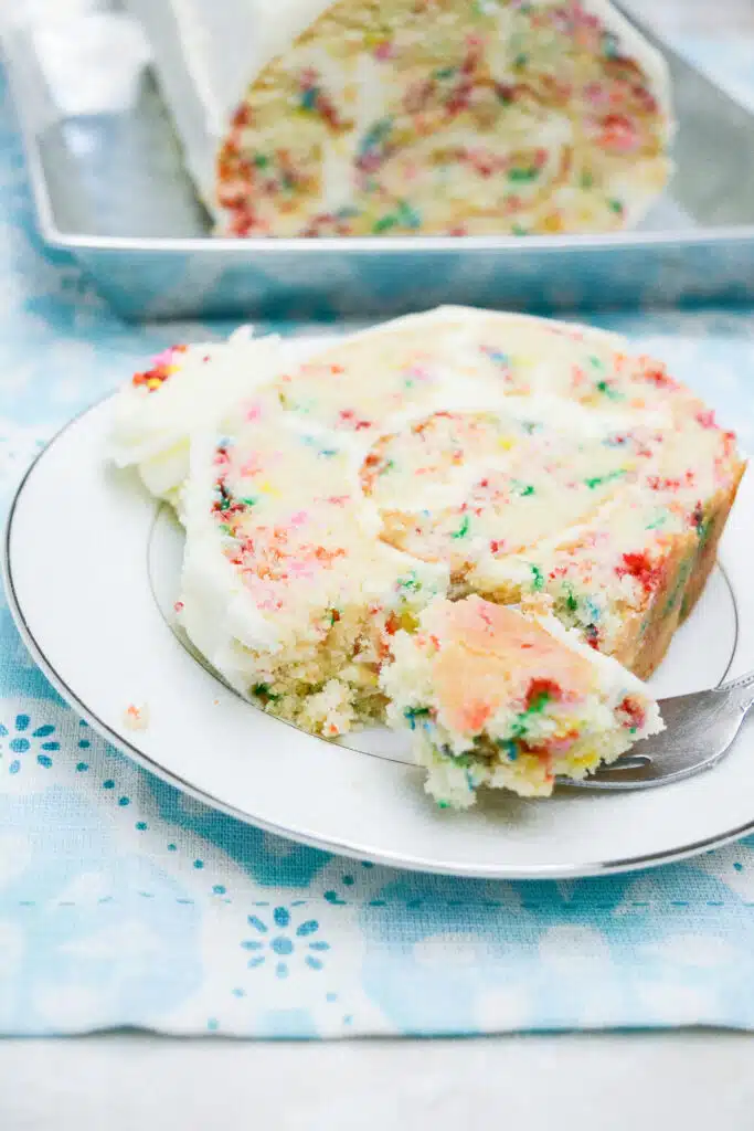 Easy From Scratch Confetti Birthday Cake Roll Recipe with Cream Cheese Frosting 34 A slice of the Delightful Funfetti Birthday Cake Roll on a white dessert plate, with a fork in it having cut through a piece of the cake.