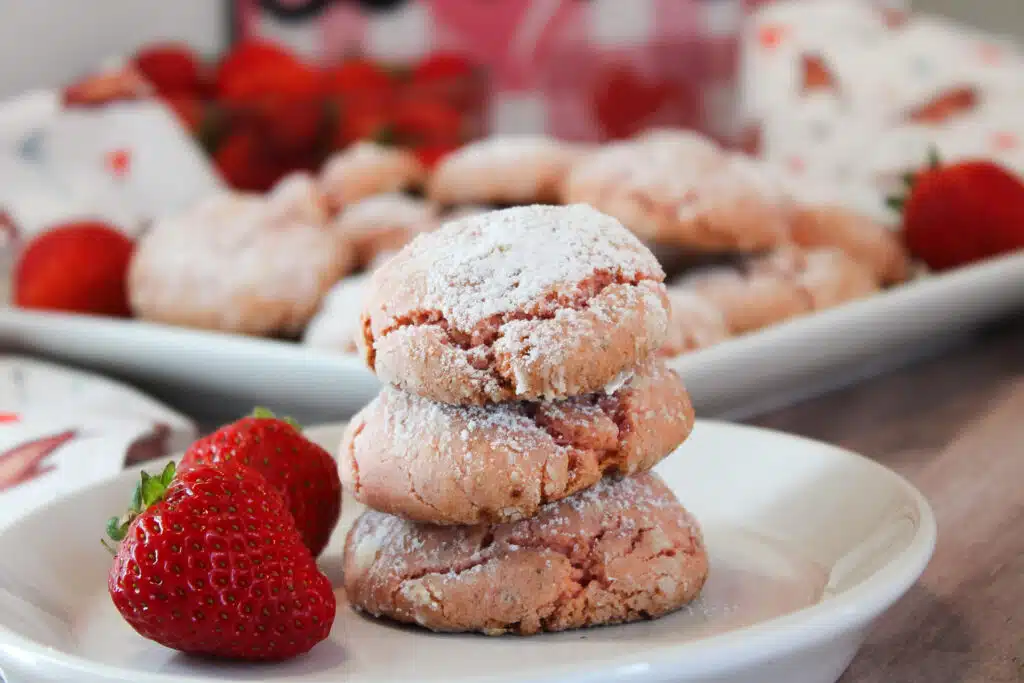 The Easiest Strawberry Cake Mix Crinkle Cookies (4 Ingredients + Vegan!) 2 Three 4-Ingredient Vegan Strawberry Cake Mix Crinkle Cookies, stacked on top of each other on a white plate with two whole fresh strawberries garnishing the plate.