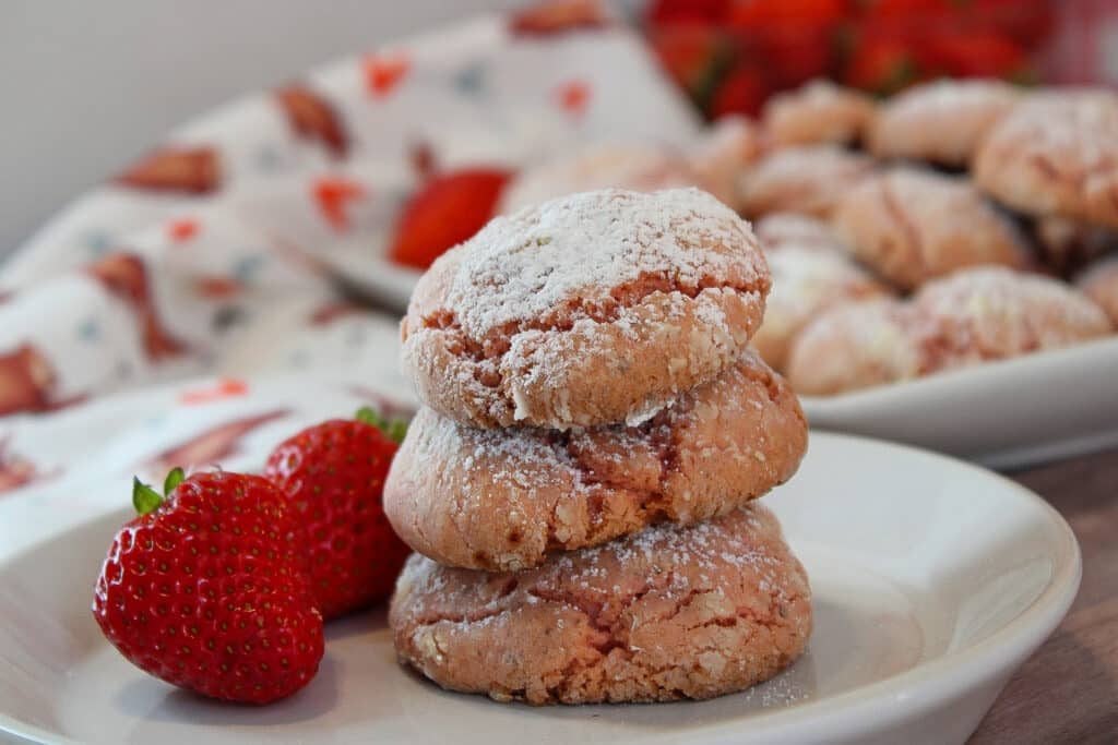 Three Vegan Strawberry Cake Mix Crinkle Cookies, stacked on top of each other on a white plate with two whole fresh strawberries garnishing the plate.