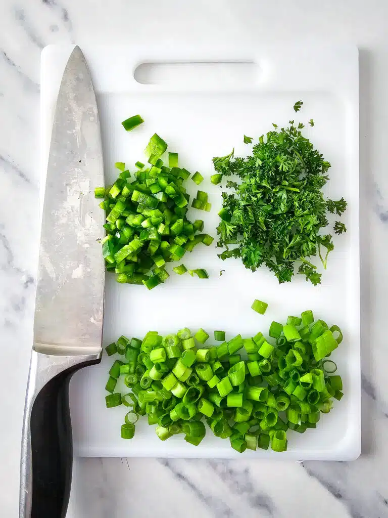 Flat lay shot of a white chopping board, with chopped jalapenos, green onions, and parsley on it. 