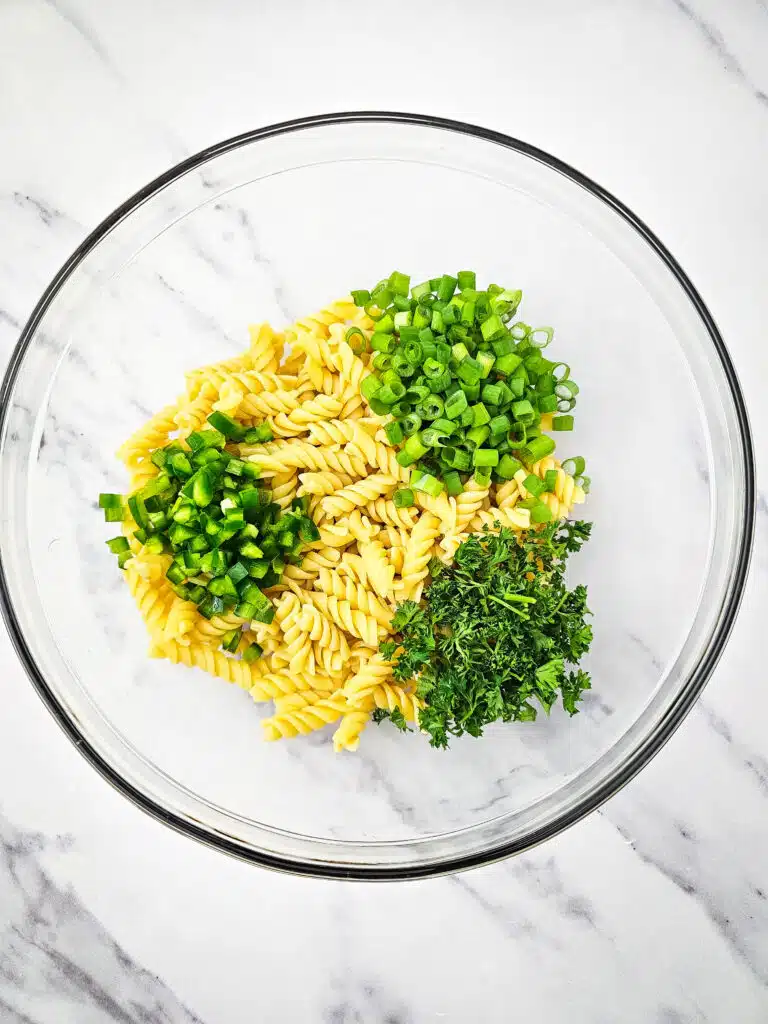 A glass bowl with the cooked rotini noodles topped with chopped jalapenos, green onions, and  parsley.