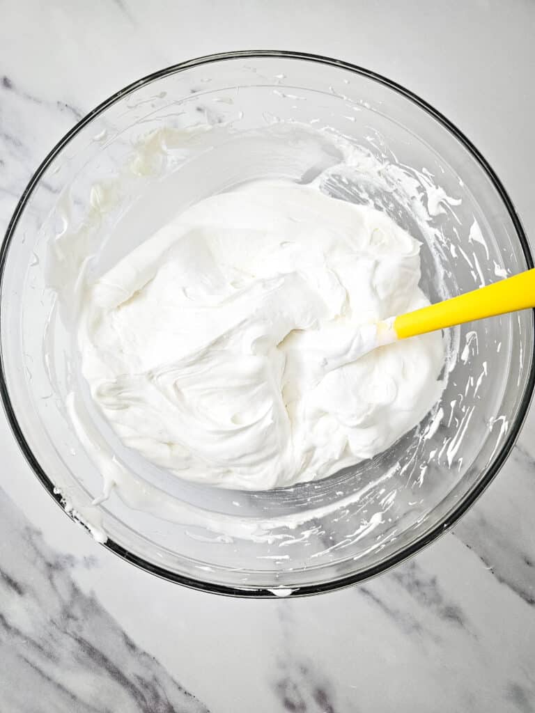 Easy 4-Ingredient Cherry Cheesecake Dip 9 Flat lay shot of a glass bowl with the cheesecake mixture in it. The cream mixture is being stirred by a yellow rubber spatula.