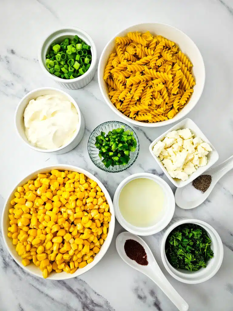 Flat lay shot of the ingredients to make the Mexican Street Corn Pasta Salad. There's drained canned corn, crumbled Cotija cheese, chopped green onions, chopped parsley, lime juice, mayonnaise, salt and pepper, rotini pasta, chopped jalapenos, and chili powder. 