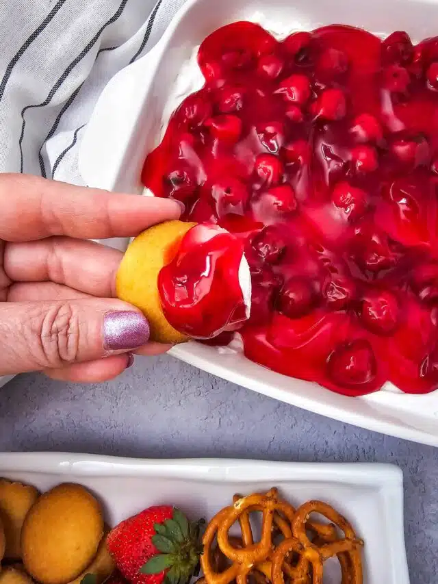 A manicured hand holding a vanilla wafer dipped in Cherry Cheesecake Dip.