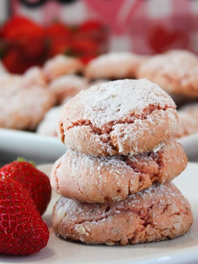 Three Vegan Strawberry Cake Mix Crinkle Cookies, stacked on top of each other on a white plate with two whole fresh strawberries garnishing the plate.
