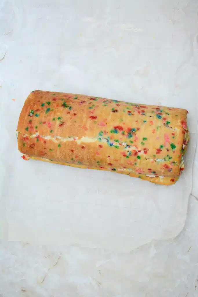 Easy From Scratch Confetti Birthday Cake Roll Recipe with Cream Cheese Frosting 27 A rolled Funfetti cake with a sheet of parchment paper underneath it.