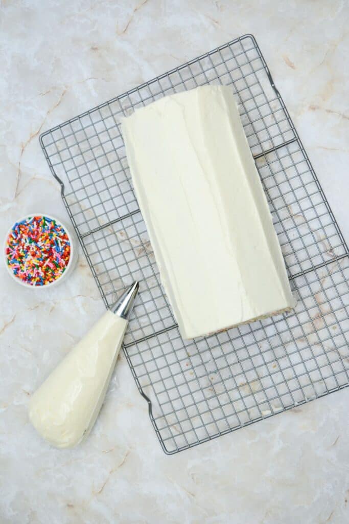 A Delightful Funfetti Birthday Cake Roll on a wire rack with a piping bag with frosting and a small colorful sprinkle beside the cake roll.
