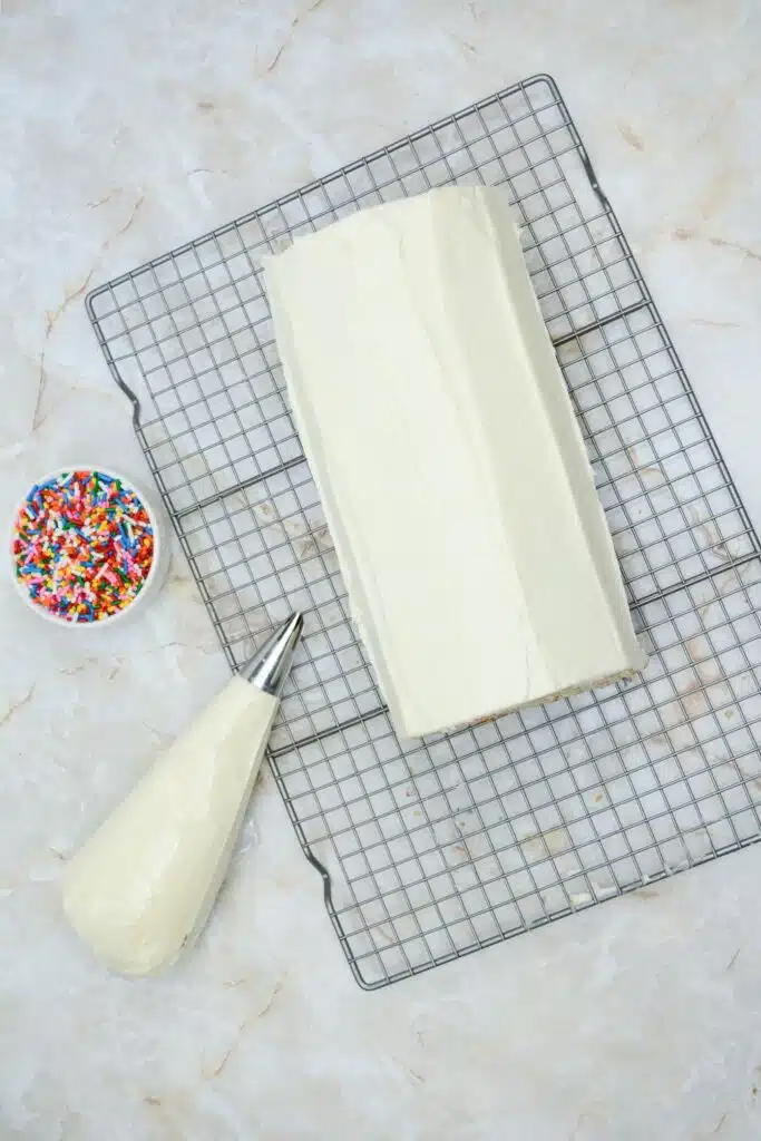 Easy From Scratch Confetti Birthday Cake Roll Recipe with Cream Cheese Frosting 30 A Delightful Funfetti Birthday Cake Roll on a wire rack with a piping bag with frosting and a small colorful sprinkle beside the cake roll.