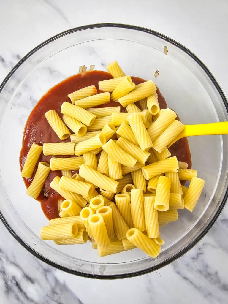 A glass bowl with tomato sauce and cooked rigatoni pasta in it.