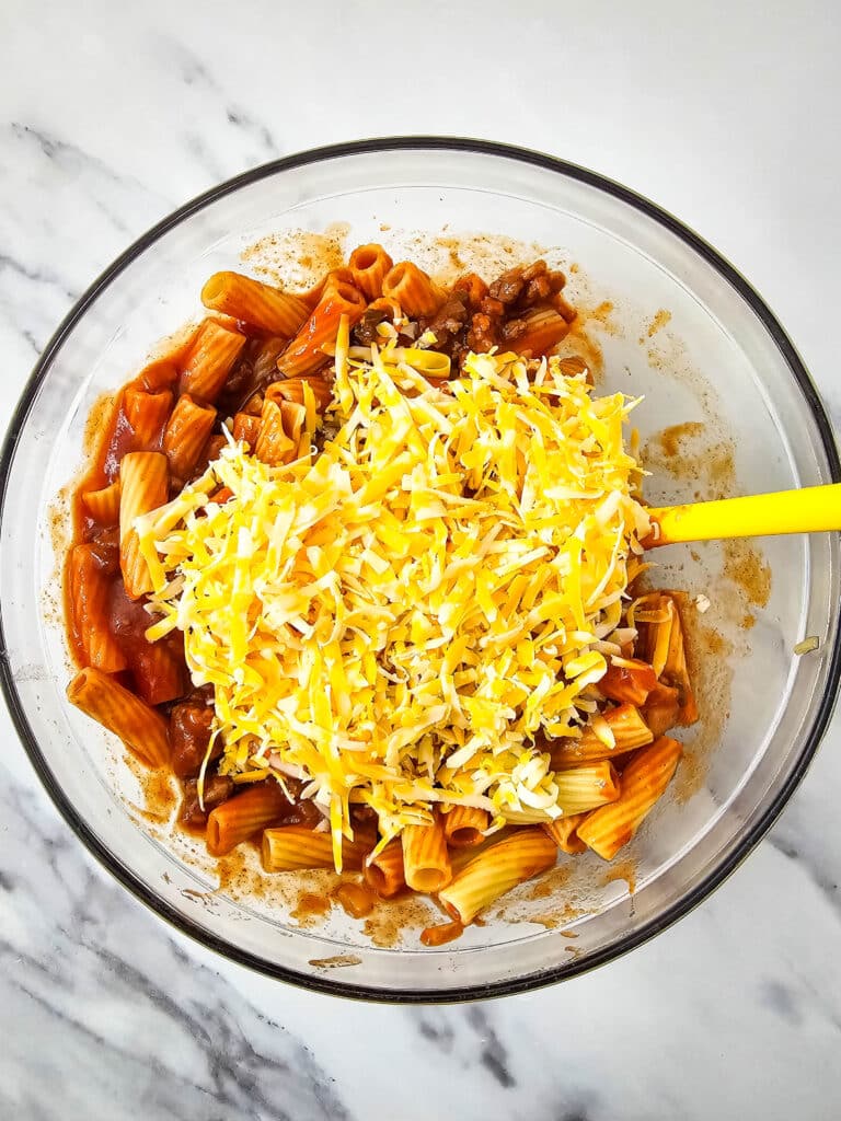 A glass bowl with pasta mixture topped with shredded cheese in it.