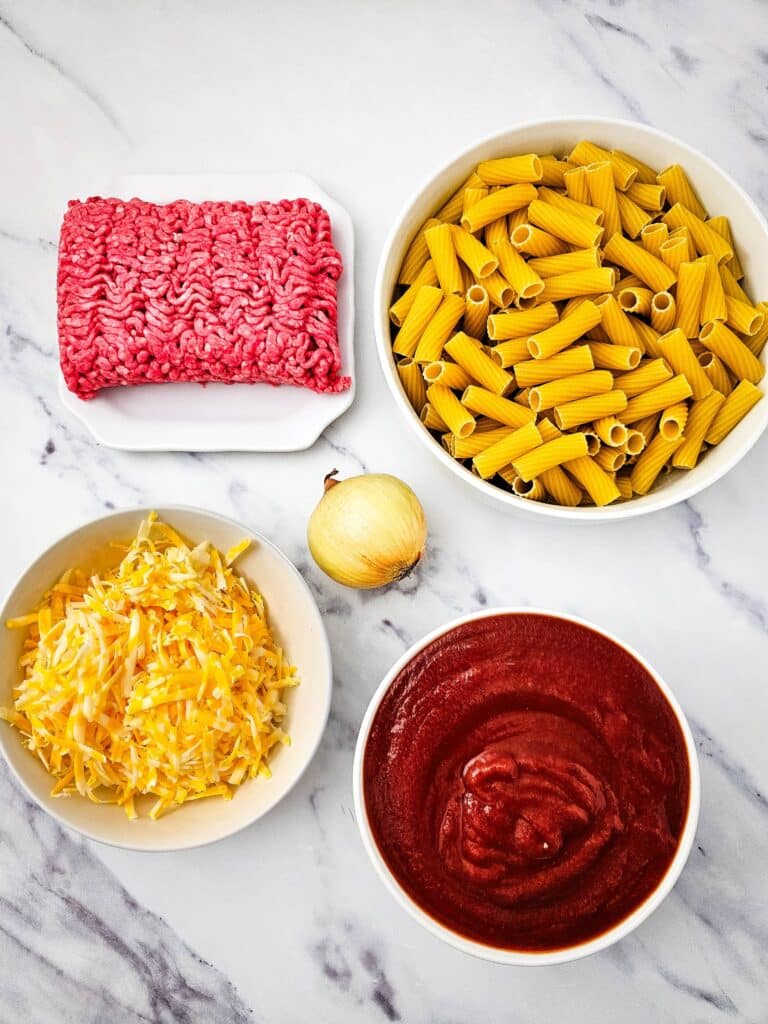 Venison Sloppy Joe Casserole Recipe 5 Flat lay shot of ingredients such as tomato sauce, shredded cheese, a yellow onion, ground venison, and uncooked rigatoni pasta. Each ingredient is on a separate white bowl and placed on a marble surface.