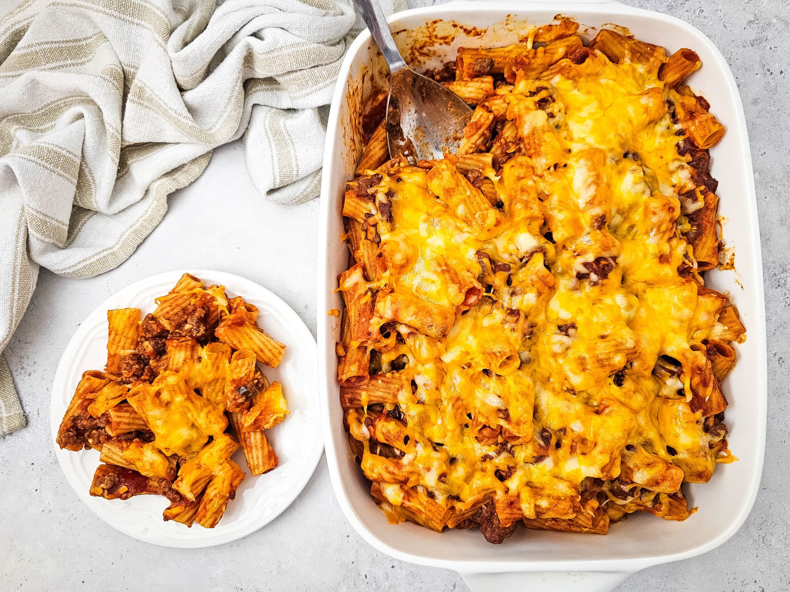 Flat lay shot of the finished Comforting and Family-Friendly Venison Sloppy Joe Casserole in a white baking dish with a serving a of it on a white plate beside the baking dish.
