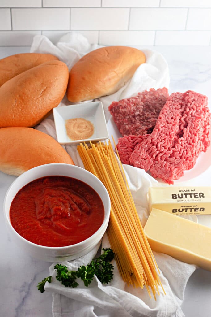 Venison Spaghetti Bread (A Family Favorite!) 6 Ingredients for the Venison Spaghetti Bread such as dry spaghetti, tomato sauce in a white bowl, half a stick of butter, ground venison on parchment paper, garlic powder, hoagie rolls.