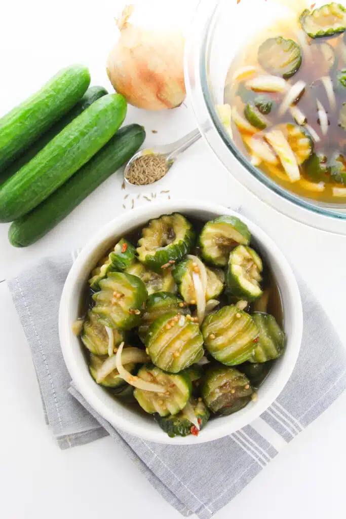 A bowl of Easy Refrigerator Bread and Butter Pickles surrounded by the ingredients and glass bowl with brine.