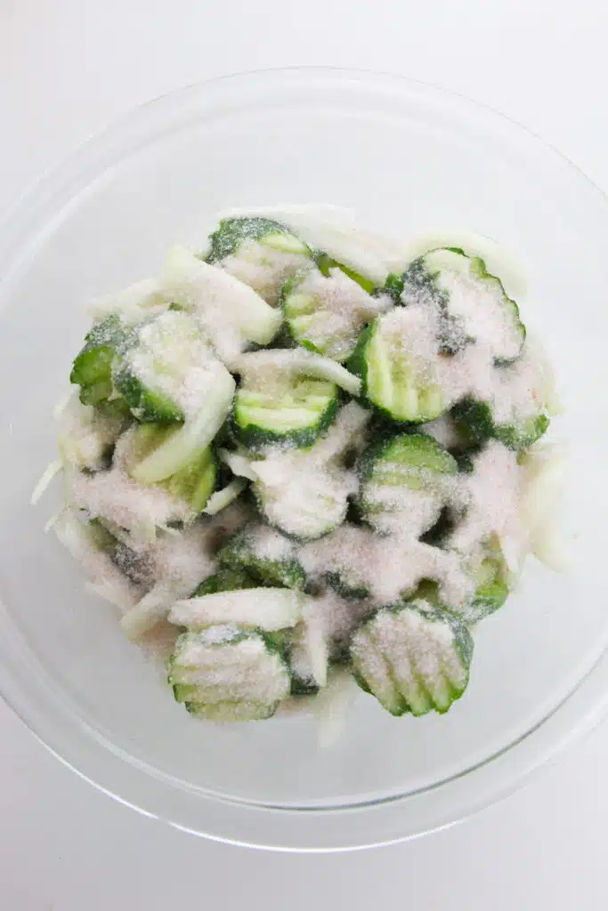 A generous amount of salt added to the glass bowl with sliced cucumbers and onions.