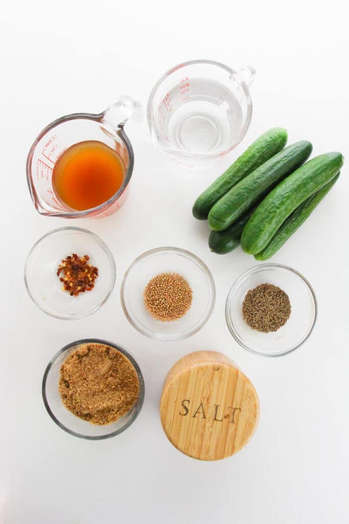 Easy Refrigerator Bread and Butter Pickles 4 Flat lay shot of the ingredients for the Bread and Butter Pickles. There's a small pile of cucumbers, a measuring jug with apple cider vinegar, another jug with distilled vinegar, red pepper flakes, mustard seeds, and coriander seeds, in separate glass bowl, another glass bowl with brown sugar and a container with salt. All ingredients are on a white surface.