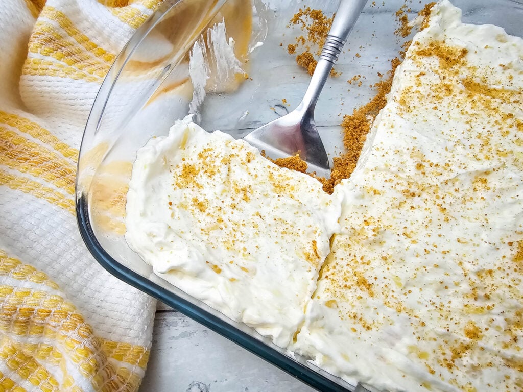 No Bake Pineapple Dream Dessert 2 A glass baking dish with a creamy dessert topped with graham cracker crumbs, a serving has been removed with a metal spatula. A yellow and white towel is beside the dish on a light surface.