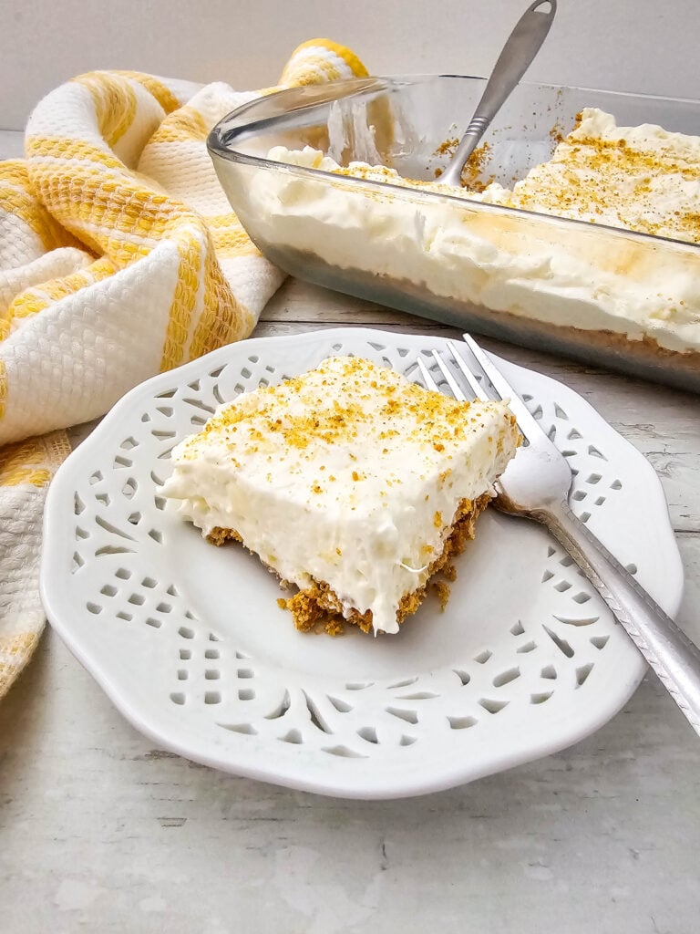 No Bake Pineapple Dream Dessert 3 A square slice of creamy dessert with a crumbly crust sits on a white decorative plate with a fork. The rest of the dessert is in a glass baking dish in the background. A yellow and white towel is nearby.