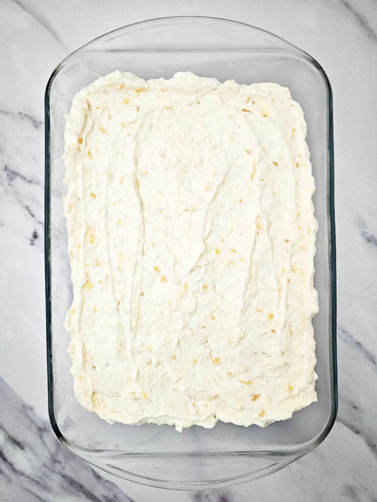 No Bake Pineapple Dream Dessert 9 A glass baking dish filled with a creamy, white mixture spread evenly, possibly a dessert or casserole, on a white marble surface. Small bits of yellow are visible throughout the mixture.