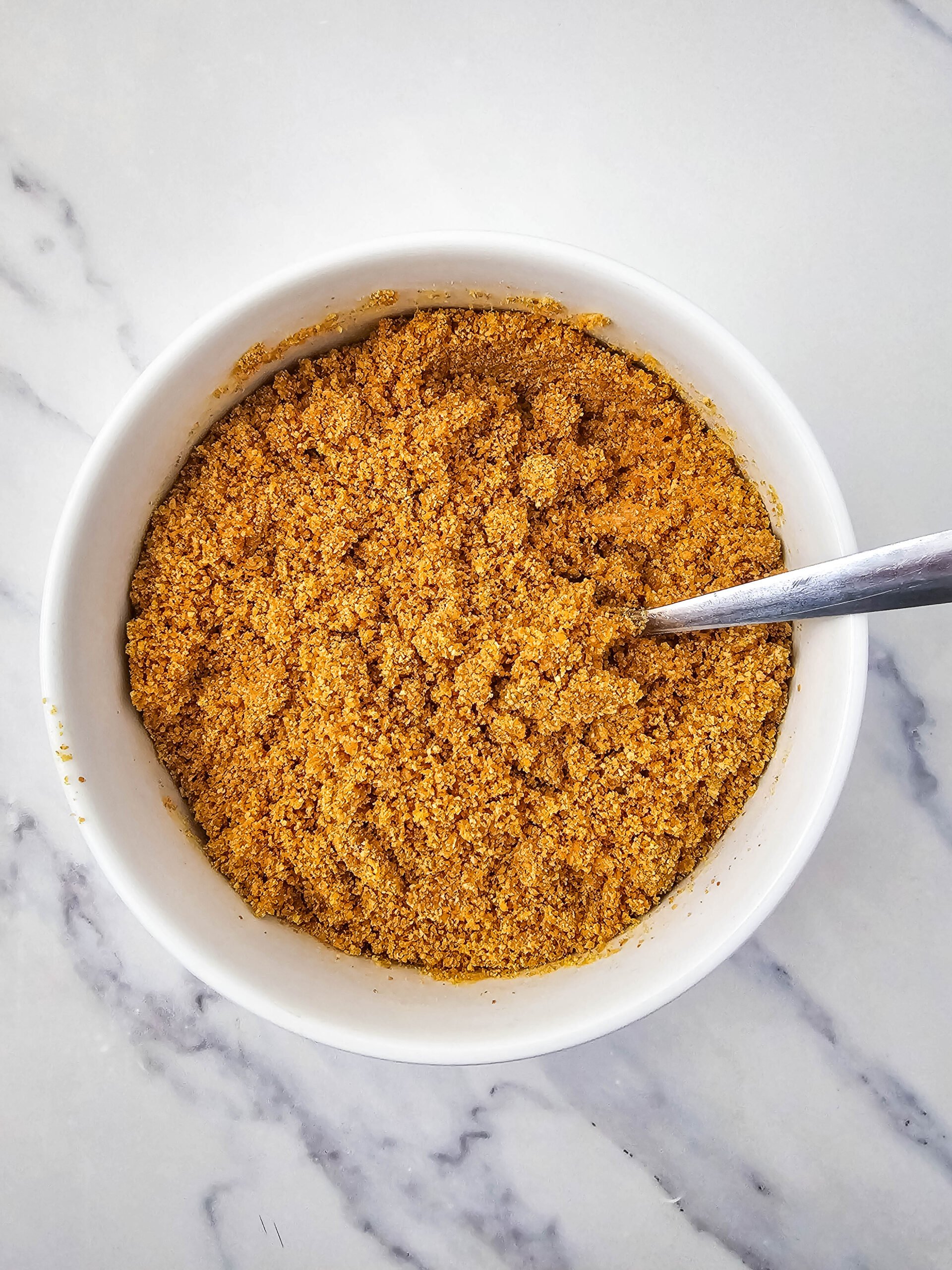 No Bake Pineapple Dream Dessert 5 A white bowl filled with a golden-brown spice mix or seasoning powder, with a metal spoon resting inside, placed on a light marble surface.