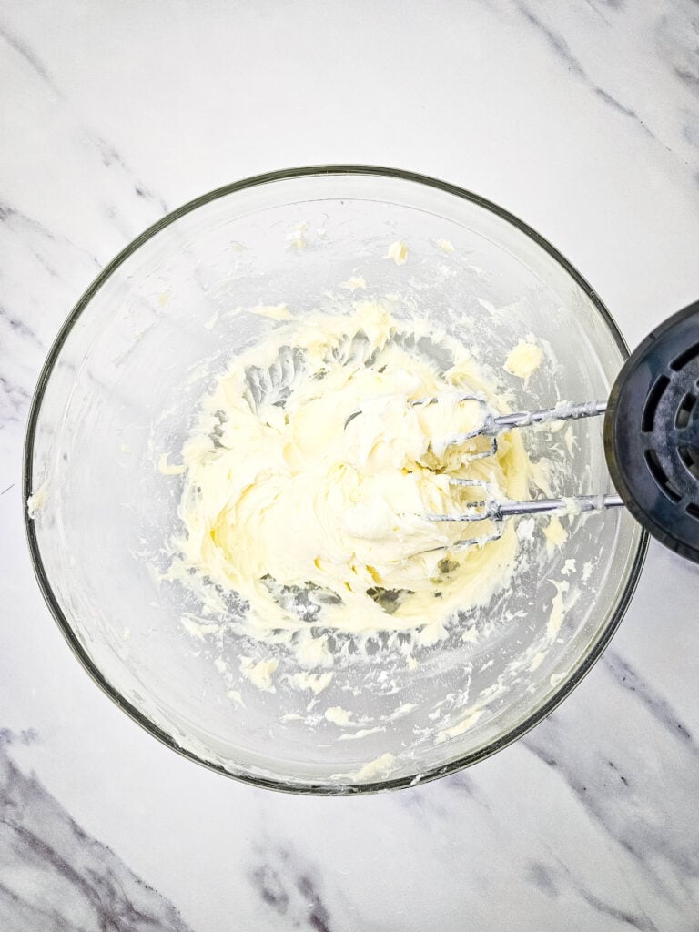 No Bake Pineapple Dream Dessert 7 A glass bowl with softened butter being mixed by an electric hand mixer on a white marble countertop.