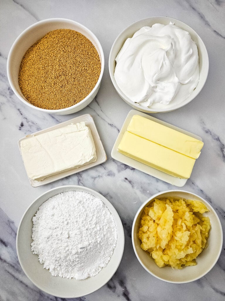 No Bake Pineapple Dream Dessert 4 Six bowls and plates on a marble surface, each containing a different ingredient: graham cracker crumbs, whipped cream, cream cheese, two sticks of butter, powdered sugar, and crushed pineapple.