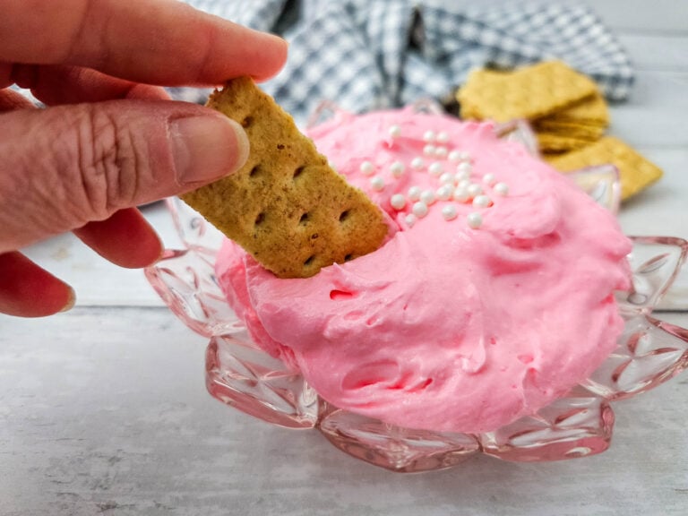 A hand dipping a piece of graham cracker into the Pink Marshmallow Fluff Dip in a glass bowl.