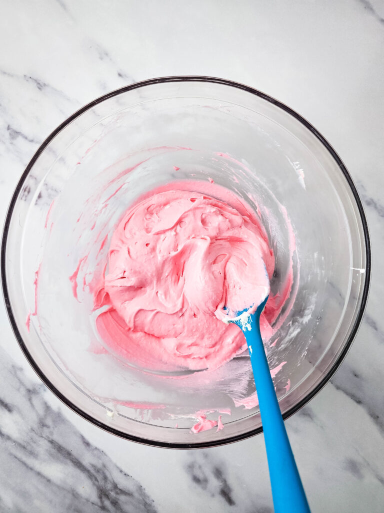 A glass bowl with pink fluff cream in it, being stirred by a blue rubber spatula.