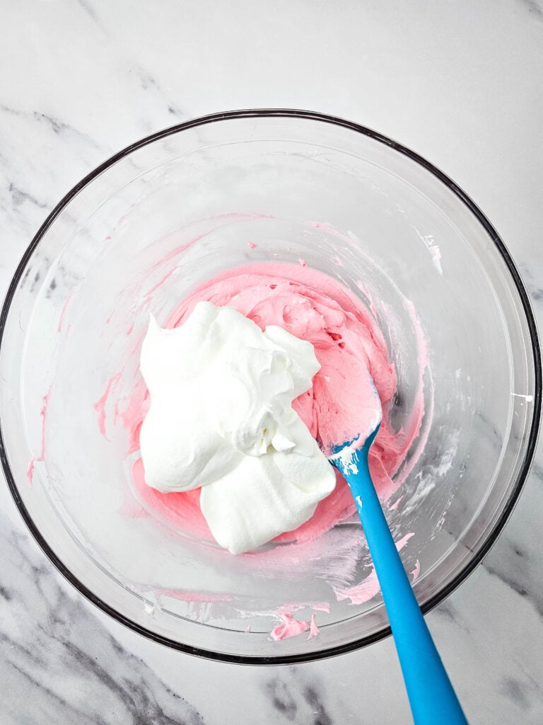 Flat lay shot of a glass bowl with pink cream topped with white whipped cream, and a blue rubber spatula, about to mix it.