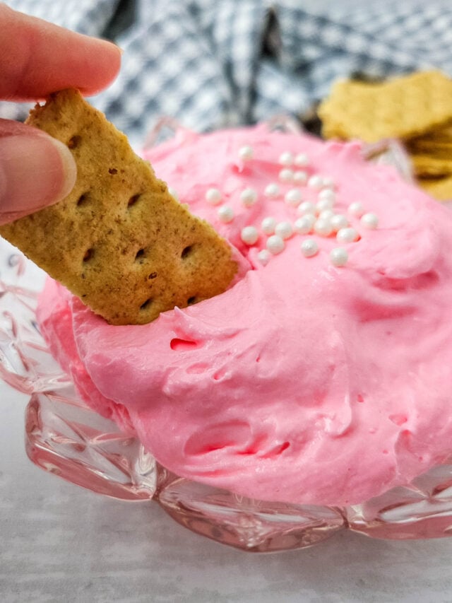 A hand dips a rectangular cracker into a bowl of pink whipped dip topped with white sprinkles; more crackers are in the background.