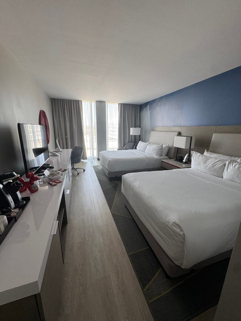 Family-Friendly Hotel in Cincinnati: Our Stay at The Summit Hotel 2 A modern hotel room with two queen beds, a desk and chair by the window, a TV, and a coffee station on a white counter. Natural light enters through tall windows with sheer curtains.
