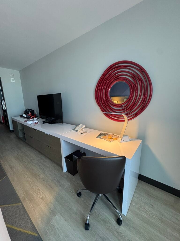 Family-Friendly Hotel in Cincinnati: Our Stay at The Summit Hotel 4 A hotel room desk with a chair, desk lamp, telephone, magazine, TV, coffee maker, and a circular red wall decoration above the desk.