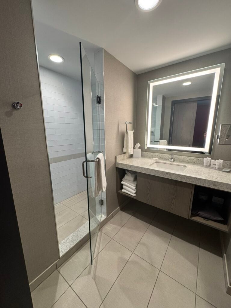 Family-Friendly Hotel in Cincinnati: Our Stay at The Summit Hotel 5 Modern hotel bathroom with a walk-in glass shower, large illuminated mirror above a sink, towels, and toiletries neatly arranged on the counter.