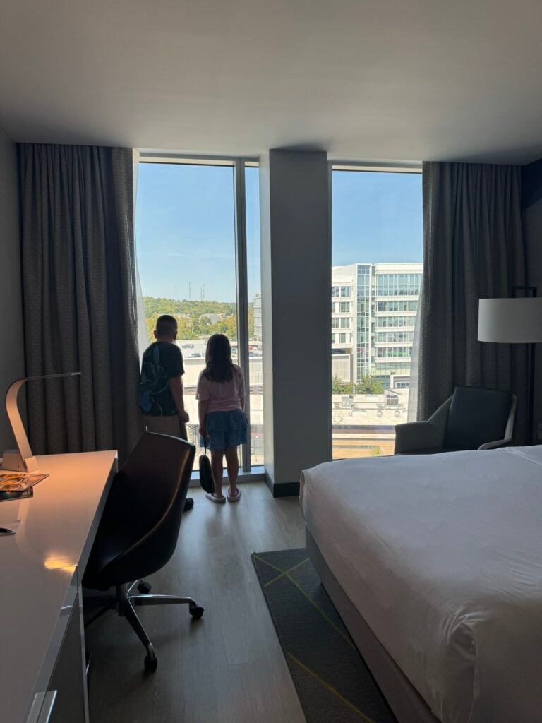 Family-Friendly Hotel in Cincinnati: Our Stay at The Summit Hotel 3 Two children stand by a large window in a hotel room, looking out at city buildings. A bed, desk, chair, and lamp are visible in the room.