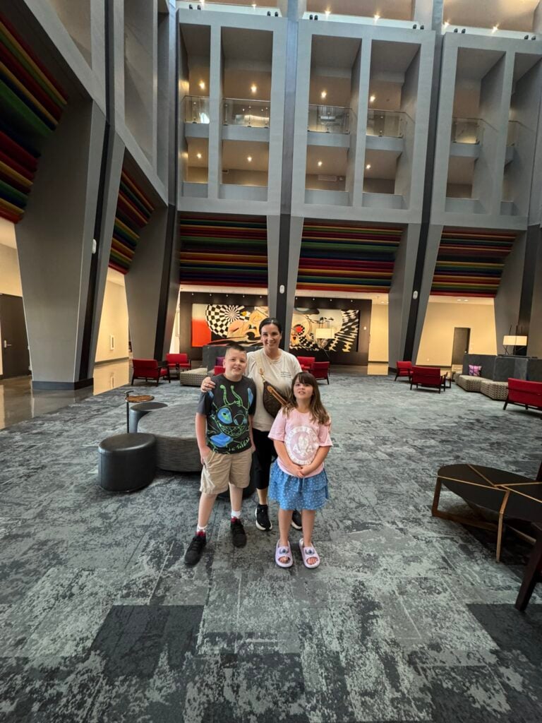 Family-Friendly Hotel in Cincinnati: Our Stay at The Summit Hotel 7 An adult and two children stand together in the center of a modern, spacious lobby with high ceilings and contemporary decor.