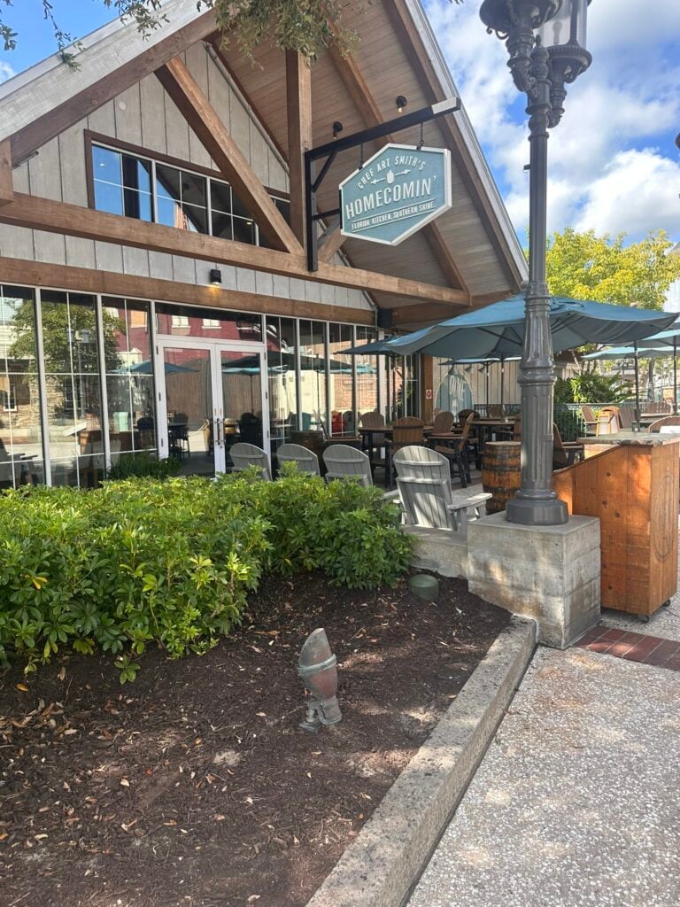 Restaurant exterior with a sign reading Homecomin’ above a patio featuring tables, chairs, umbrellas, and lush greenery under a partly cloudy sky—one of the Top 3 Must-Pick Family Friendly Restaurants at Disney Springs.