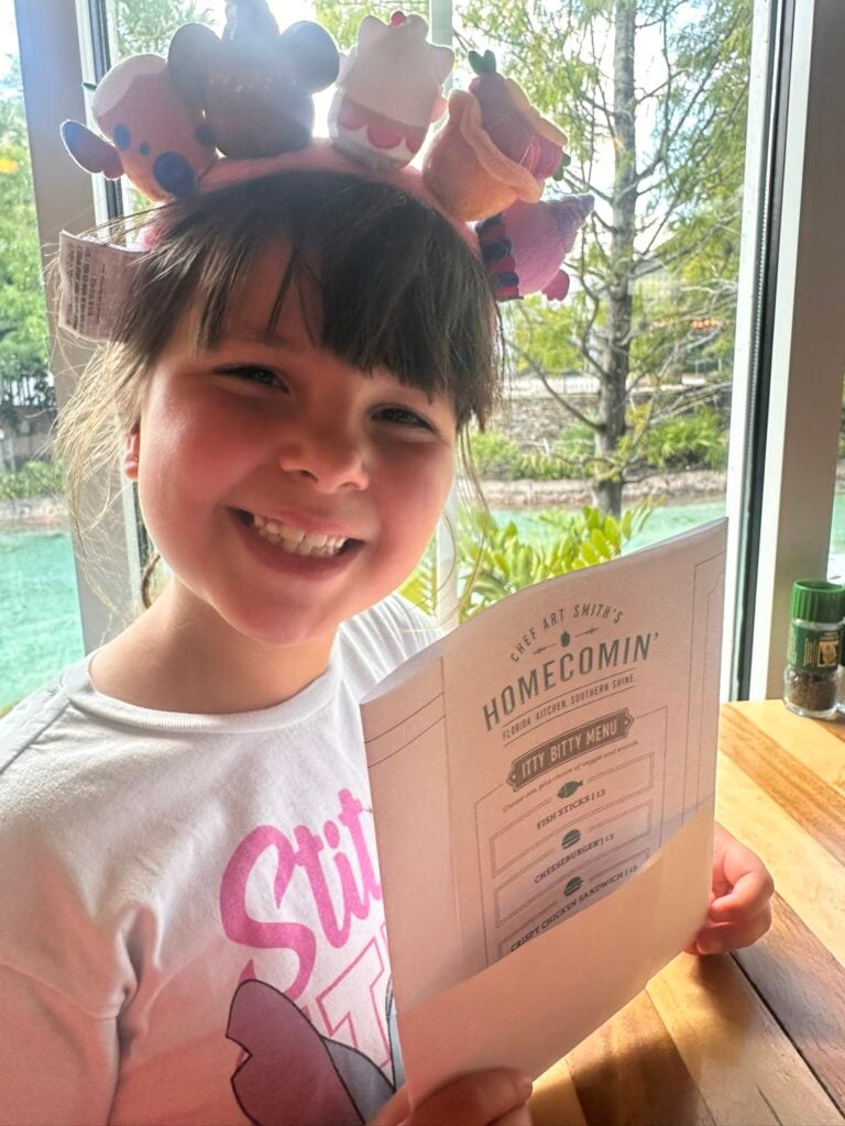 Smiling child wearing a plush animal headband holds a menu at a restaurant table by a window, enjoying the view of trees and water at one of the Top 3 Must-Pick Family Friendly Restaurants at Disney Springs.