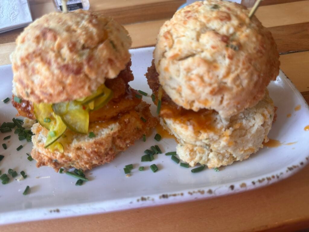 Two biscuit sliders filled with fried chicken, pickles, and sauce are served on a white plate, garnished with chopped chives—no wonder it’s featured at one of the Top 3 Must-Pick Family Friendly Restaurants at Disney Springs.