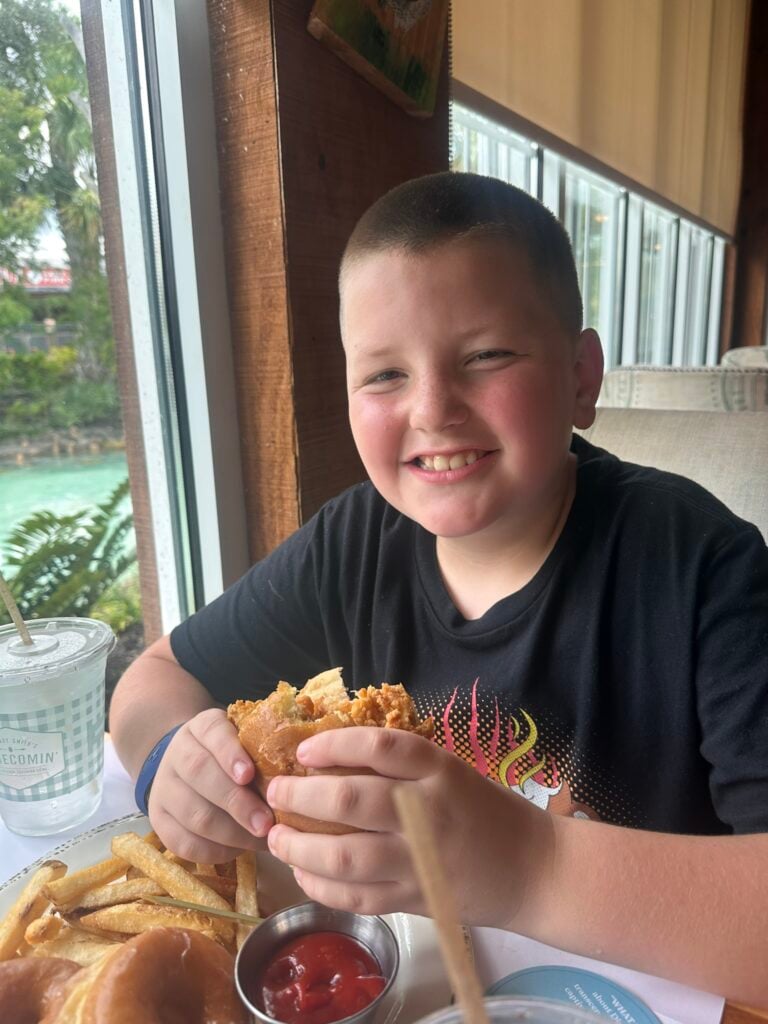 A boy with short hair sits at a table by a window, smiling while holding a sandwich. French fries, ketchup, and a drink are on the table—capturing a moment at one of the Top 3 Must-Pick Family Friendly Restaurants at Disney Springs.