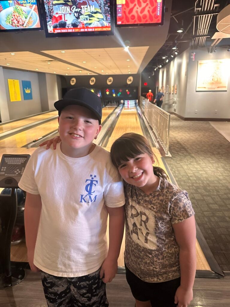 Two children stand side by side and smile at a bowling alley, one of the Top 3 Must-Pick Family Friendly Restaurants at Disney Springs, with bowling lanes and screens visible in the background.