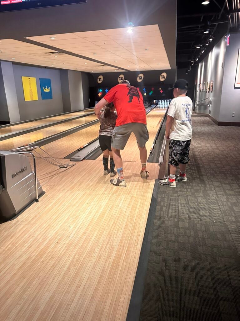 An adult helps a child bowl while another child stands nearby in a bowling alley, just steps away from the Top 3 Must-Pick Family Friendly Restaurants at Disney Springs, with pins visible at the end of the lane.