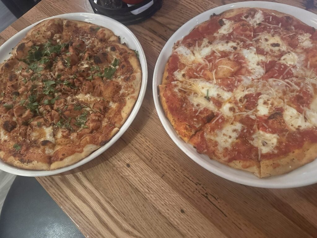 Two pizzas on white plates sit on a wooden table—one topped with herbs and meat, the other with tomato sauce and melted cheese—perfect for sharing at one of the Top 3 Must-Pick Family Friendly Restaurants at Disney Springs.