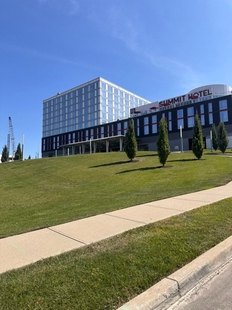 A modern hotel building labeled Summit Hotel sits atop a grassy hill under a clear blue sky, with a sidewalk and young trees in the foreground.